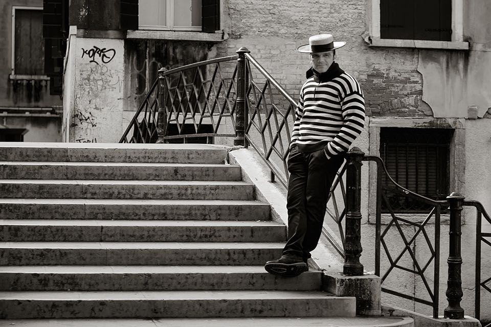 Das Portraitfoto zeigt einen Gondoliere mit Hut und gestreiftem Hemd auf einer Bruecke in Venedig